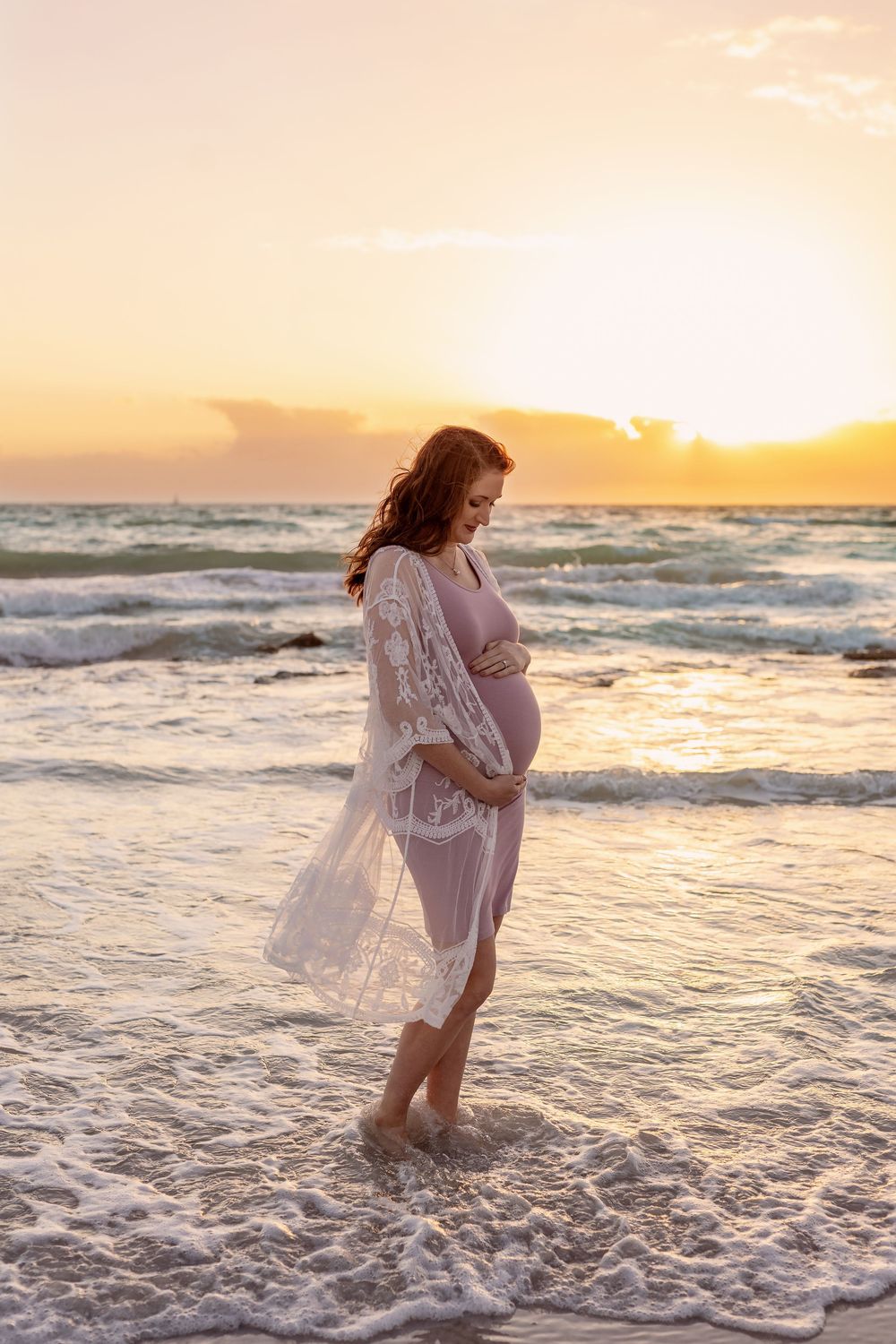 Pregnant woman in pink dress and lace robe standing in ocean waves at golden sunset, cradling baby bump.