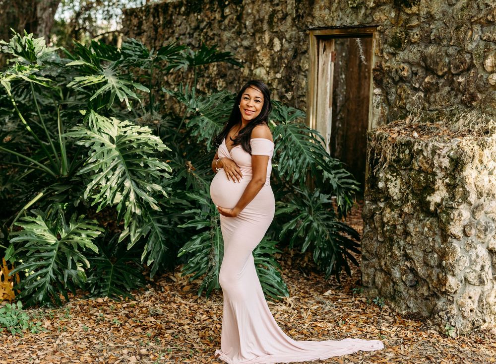 Pregnant woman in blush pink off-shoulder gown poses gracefully outdoors near tropical foliage and rustic stone wall.
