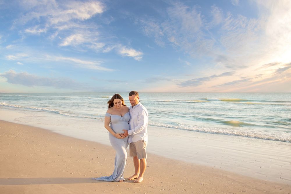 Pregnant couple posing on a sandy beach at sunset, with turquoise ocean waves and a blue sky behind them.
