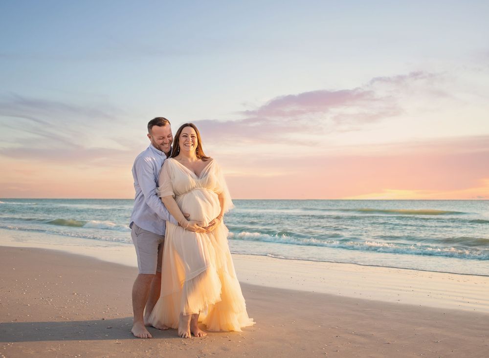 Couple poses on beach at sunset, woman in flowing gown cradling baby bump, soft pastel sky and ocean backdrop.