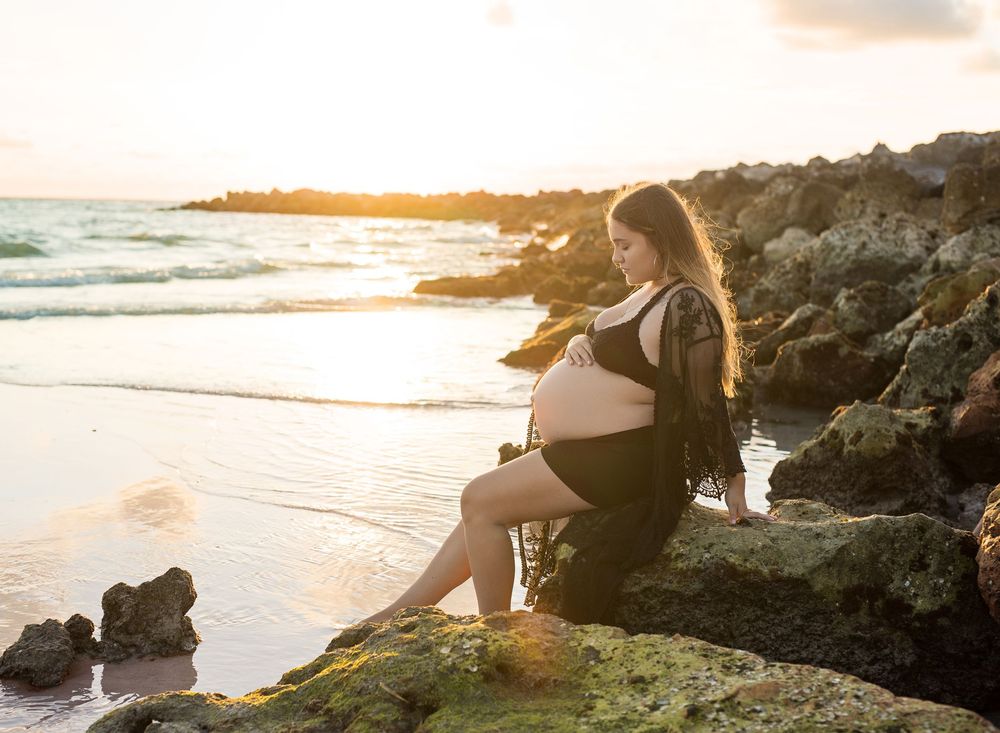 Pregnant woman in black dress sitting on rocky beach at sunset with ocean waves in background.