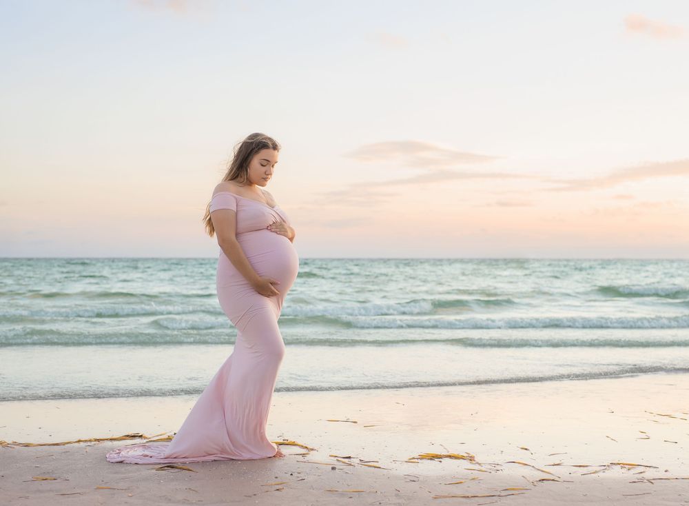 Pregnant woman in pink gown poses on beach at sunset, cradling her baby bump with ocean waves behind her.