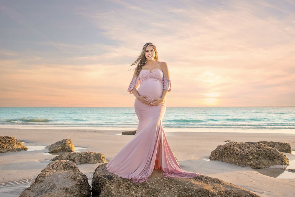 Pregnant woman in pink maternity gown posing on beach rocks at sunset with turquoise ocean waves behind her.