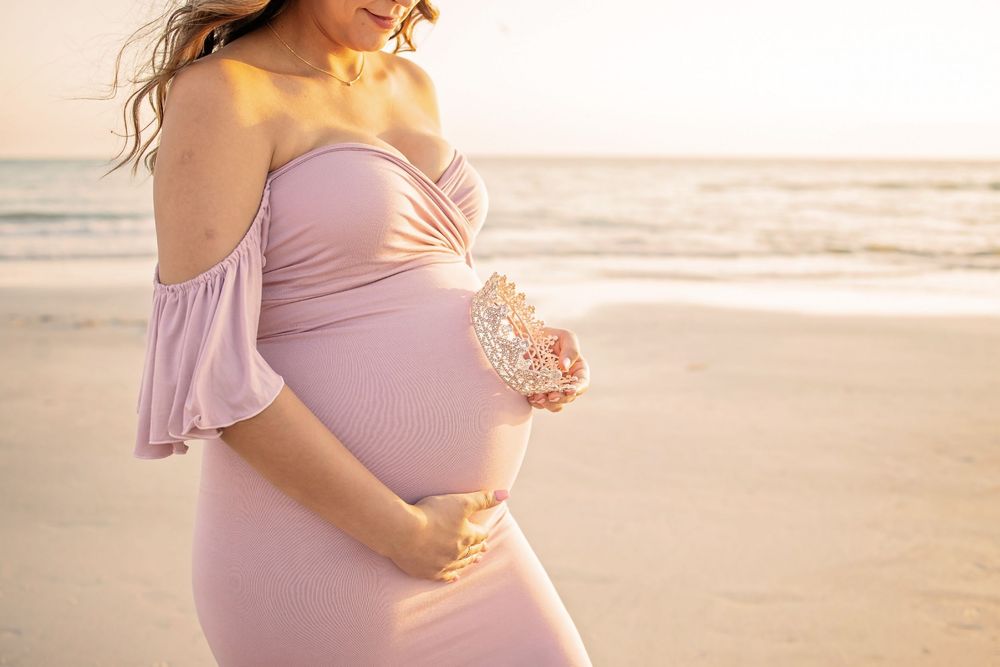 Pregnant woman in pink off-shoulder gown cradling baby bump on beach at golden hour sunset.