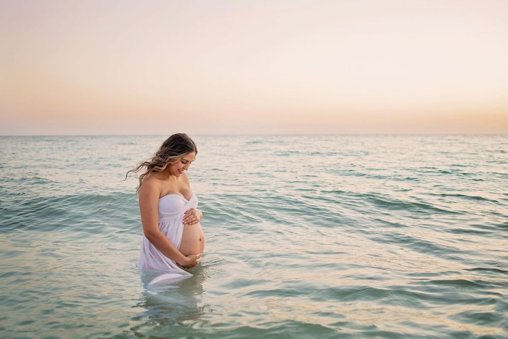 Pregnant woman in white dress standing in calm ocean water at sunset, cradling her baby bump.