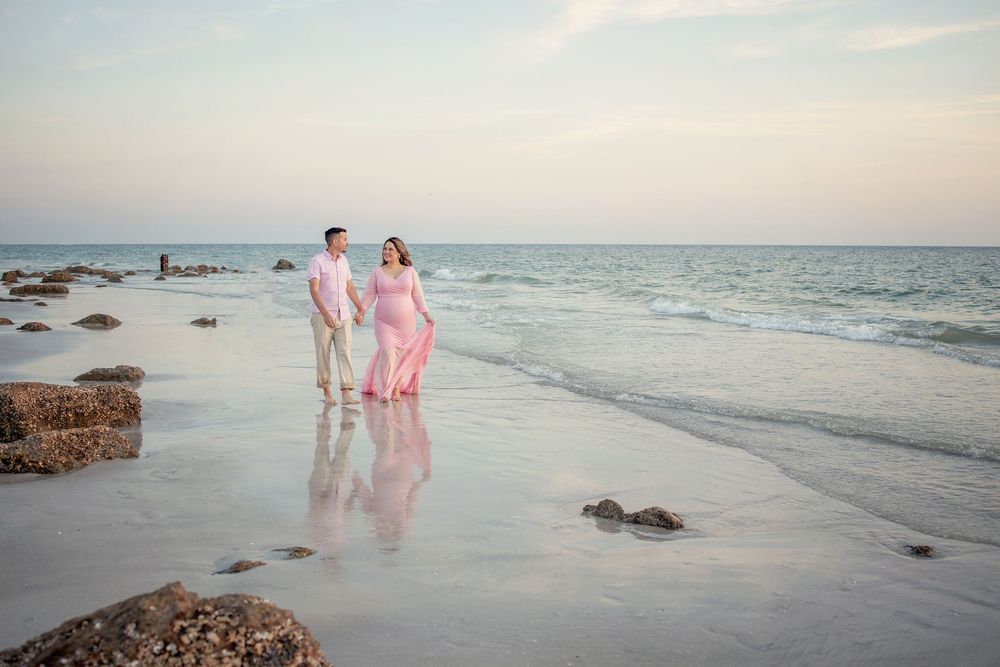 Couple walking hand-in-hand on a sandy beach at sunset, woman in pink flowing dress, calm turquoise ocean waves behind them.