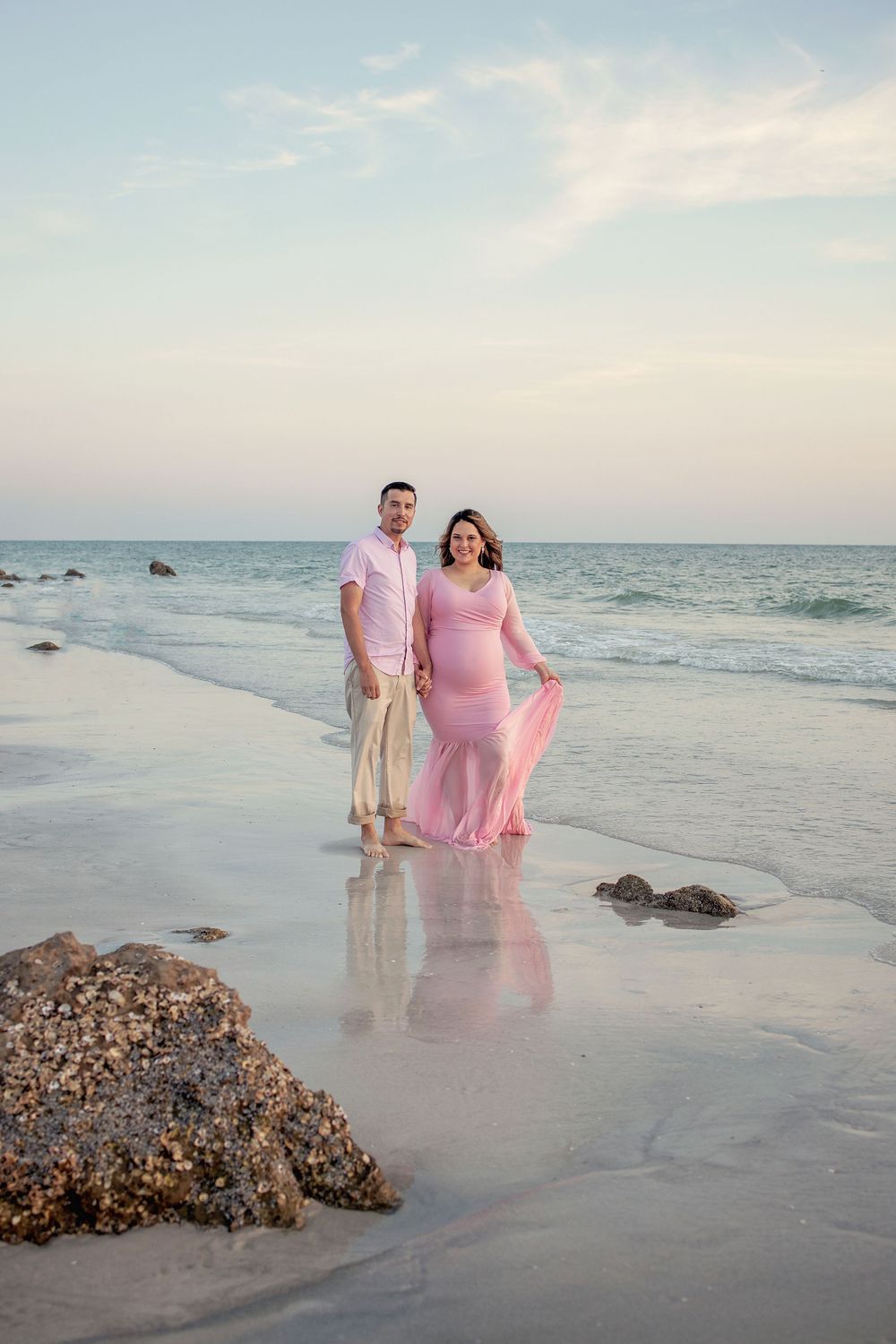 Pregnant couple in pink outfits standing on a sandy beach at sunset with ocean waves in the background.