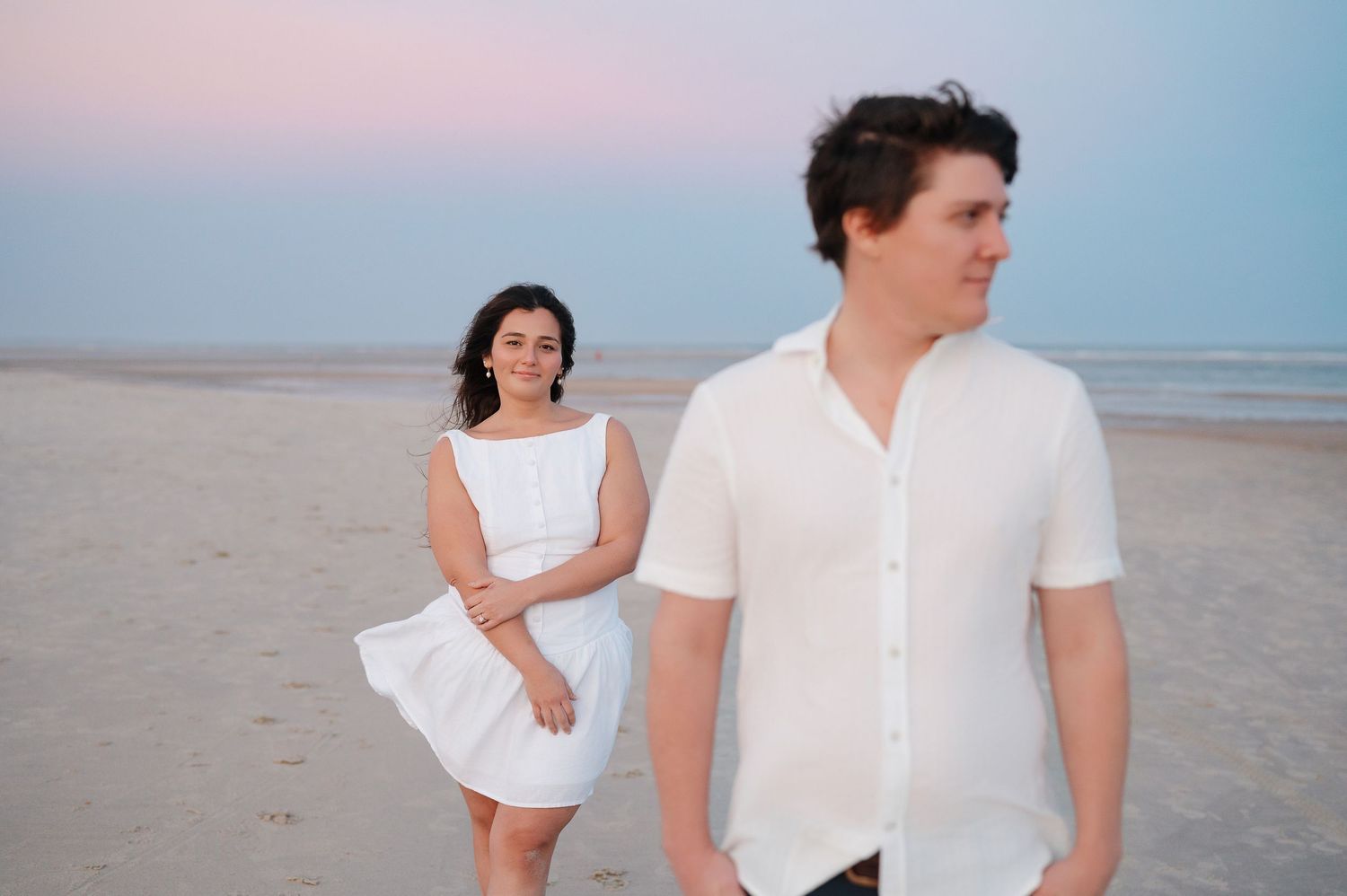 A couple in white outfits poses separately on a beach during sunset.