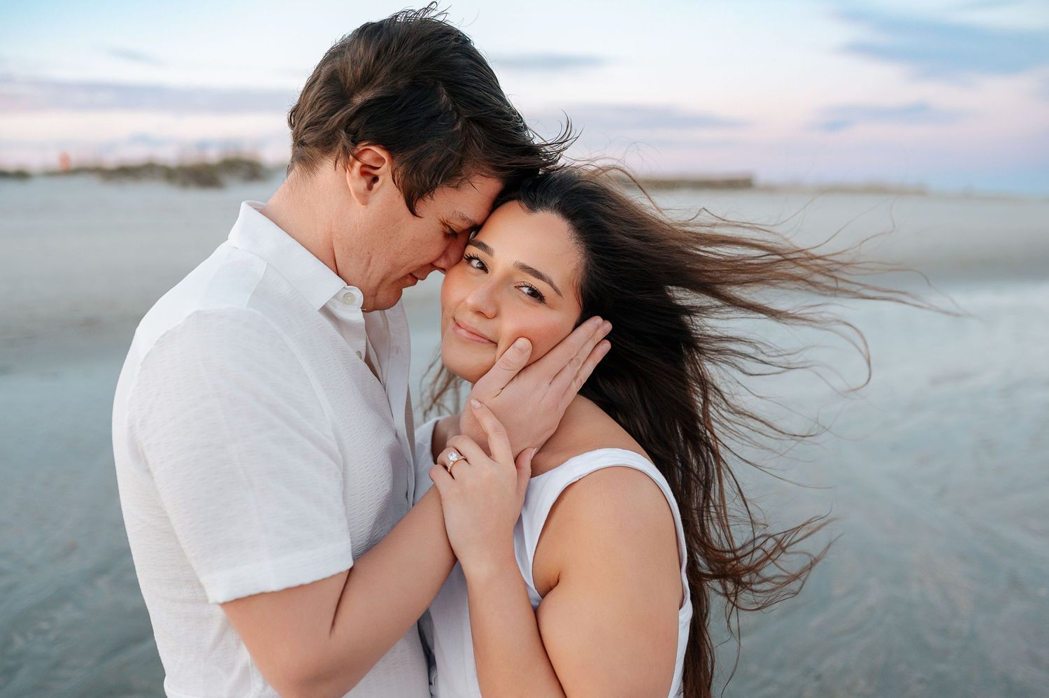 Romantic beach photoshoot at sunset with couple embracing in white and tan outfits against ocean backdrop.