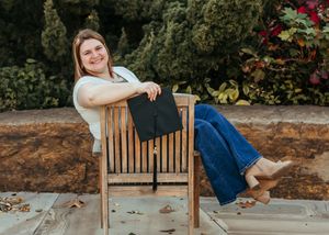 A college graduate poses with her cap, kicking back in a wooden chair with foliage in the background.