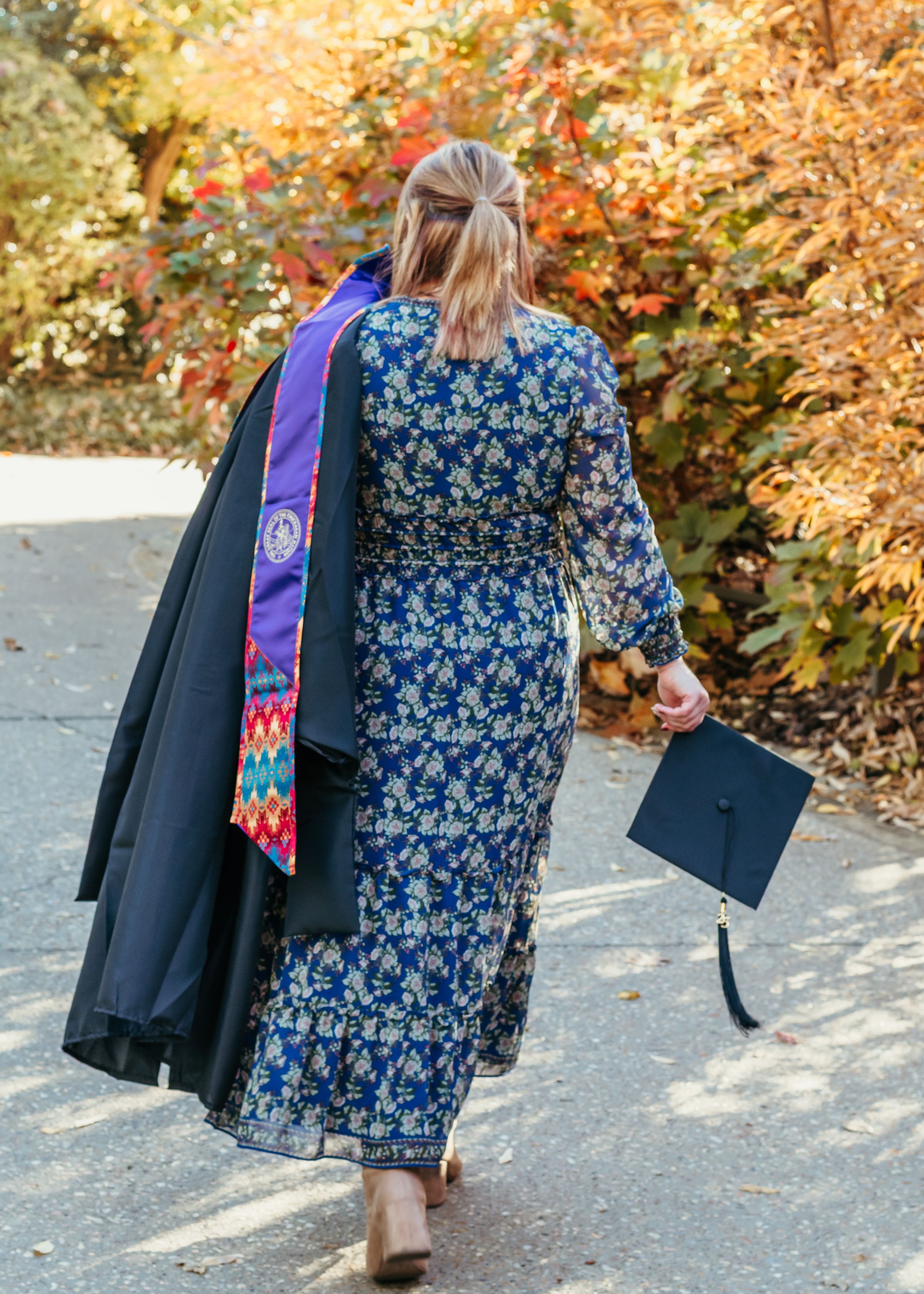 A college graduate wearing a blue floral dress carries a her cap and gown while walking outdoors in autumn scenery.