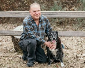 A man in plaid shirt and jeans sits on wooden steps with a black dog in a natural outdoor setting.