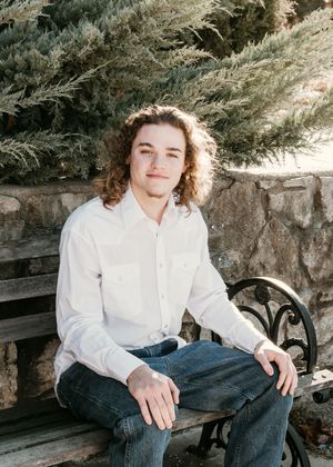 A person in a white button-down shirt and jeans sits casually on a stone bench against a rustic wall background.