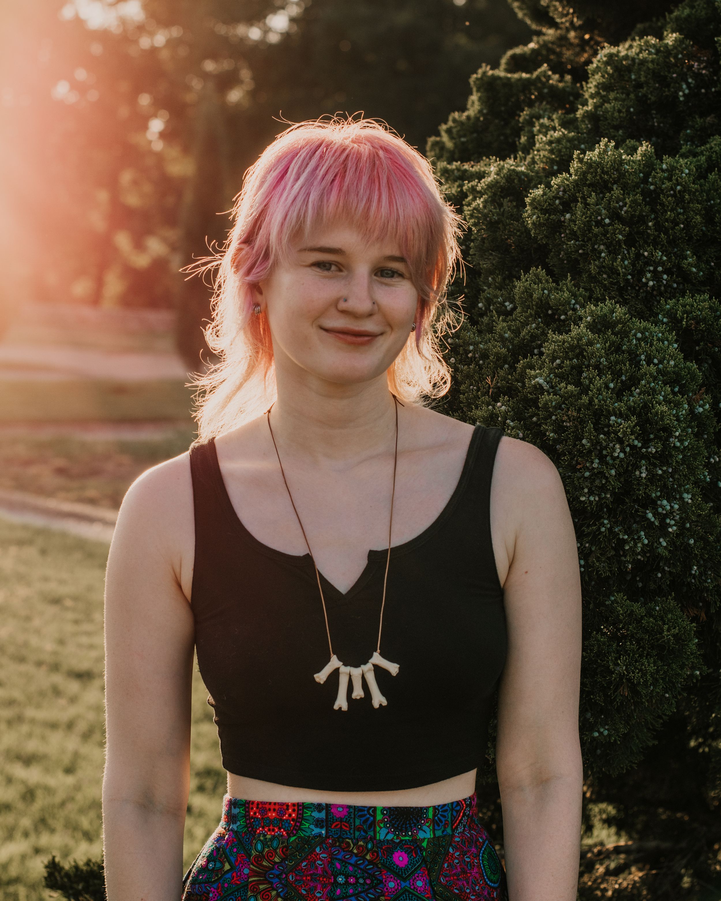 A person with pink hair wearing a black top and patterned skirt poses outdoors during golden hour sunset.