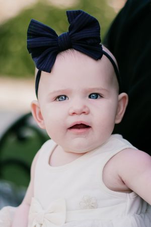 A baby wearing a navy blue bow headband and white outfit coos sweetly in an outdoor portrait series.