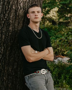 A high school senior in a black t-shirt and light jeans poses against a tree trunk in a wooded outdoor setting.
