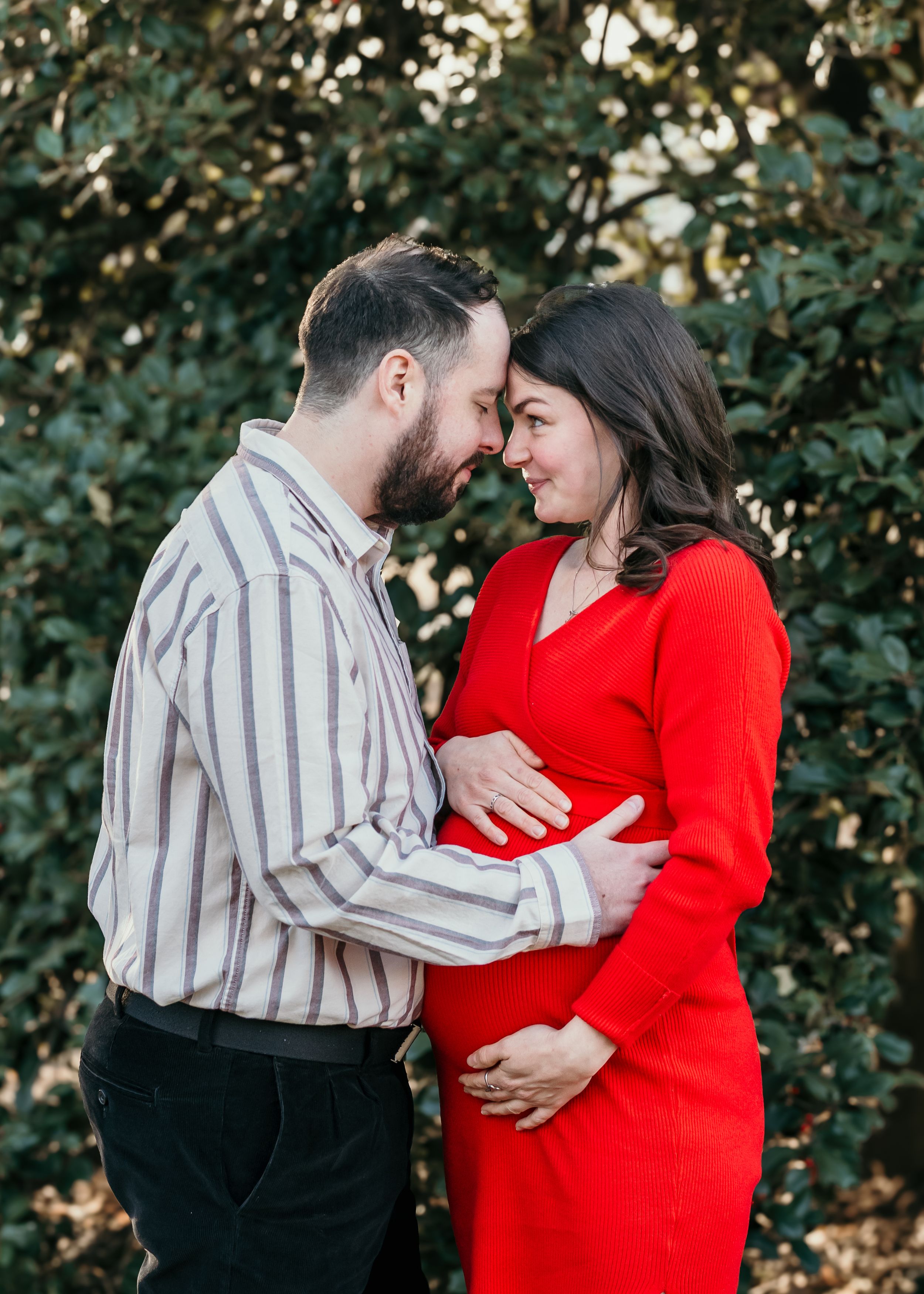 A couple in a romantic outdoor setting with green foliage, wearing a red dress and striped shirt while embracing at a maternity session.