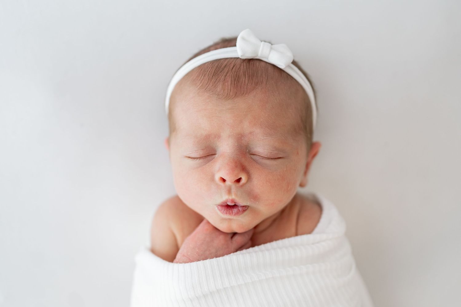 Peaceful newborn baby sleeping soundly wrapped in soft white blankets against neutral background.