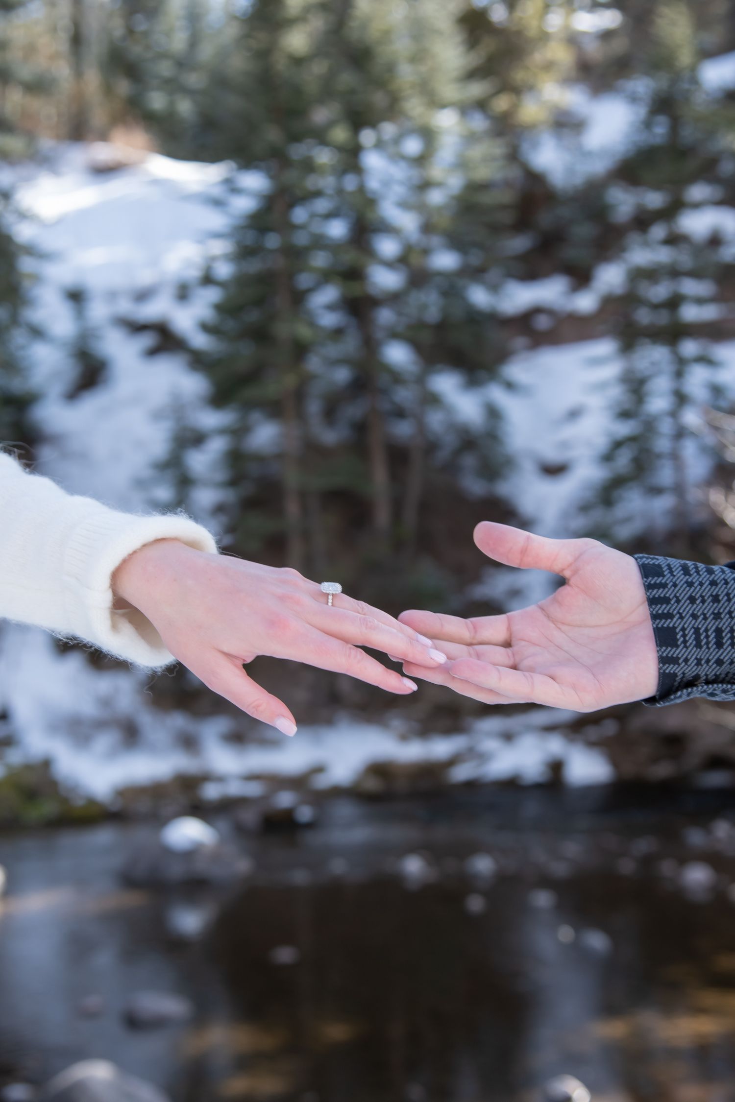 Two hands reaching towards each other against a snowy winter forest background with water after a surprise winter proposal in Vail Colorado