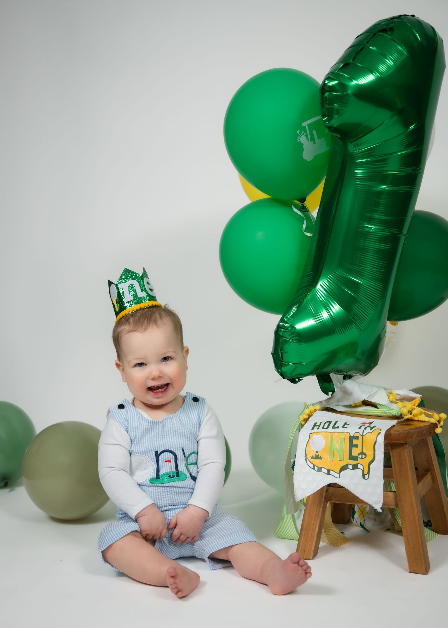 Baby wearing green crown and white onesie sits on floor with St. Patrick's Day themed green balloon decorations.