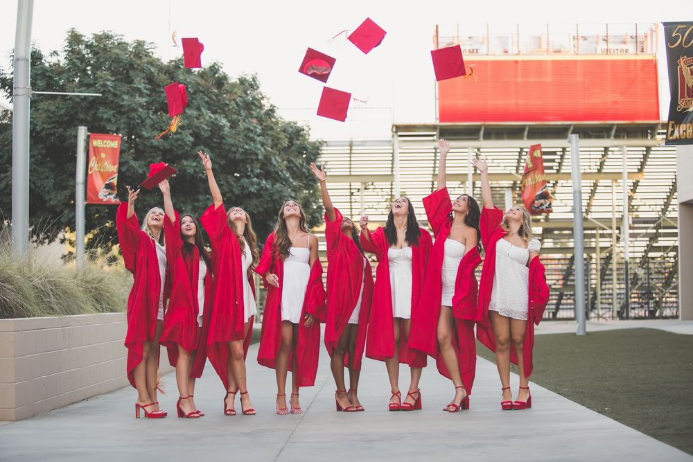 A group in red graduation gowns toss their caps in celebration on a sunny day outside a modern building.