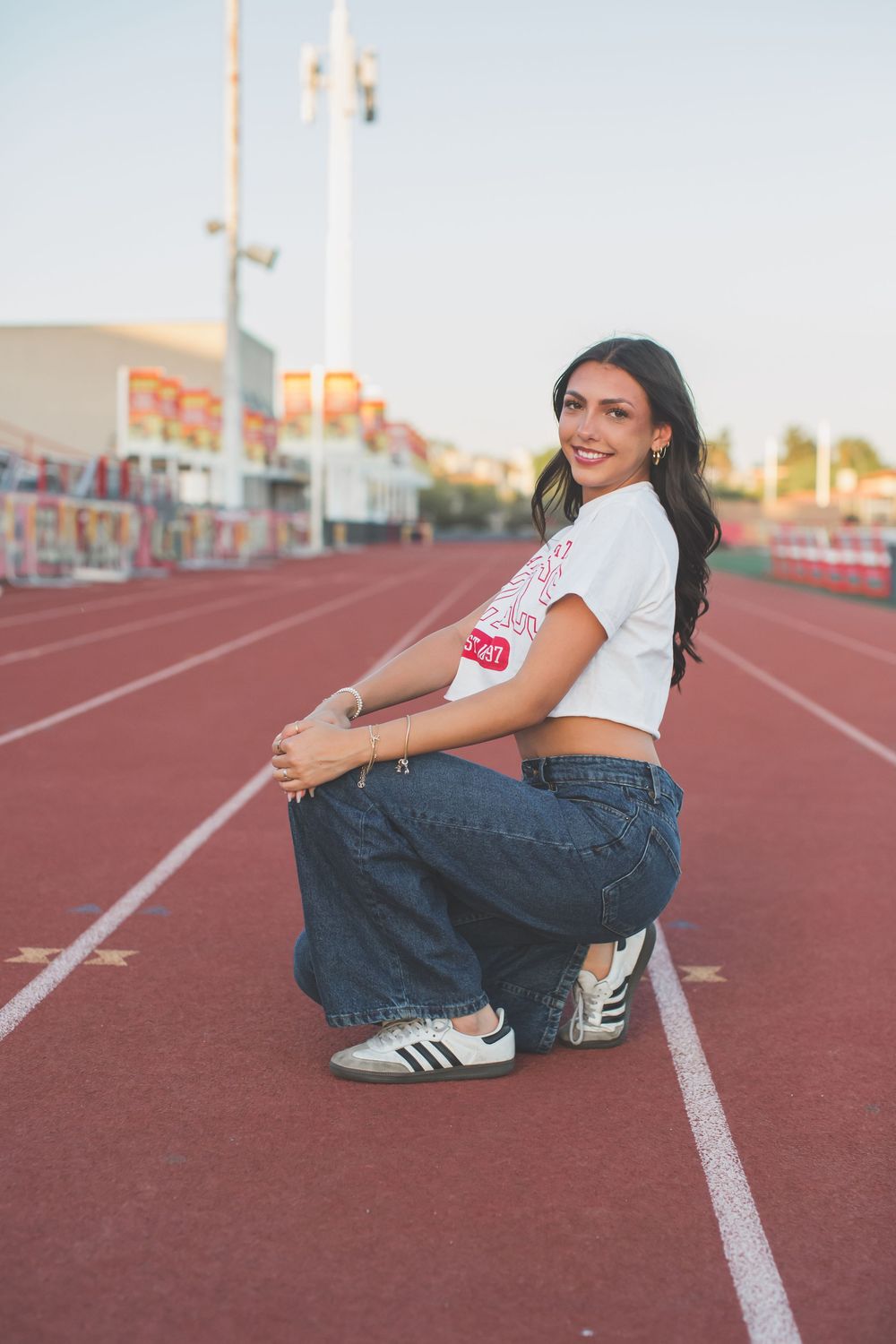 Person in casual white t-shirt and jeans poses playfully on red running track at sunset.