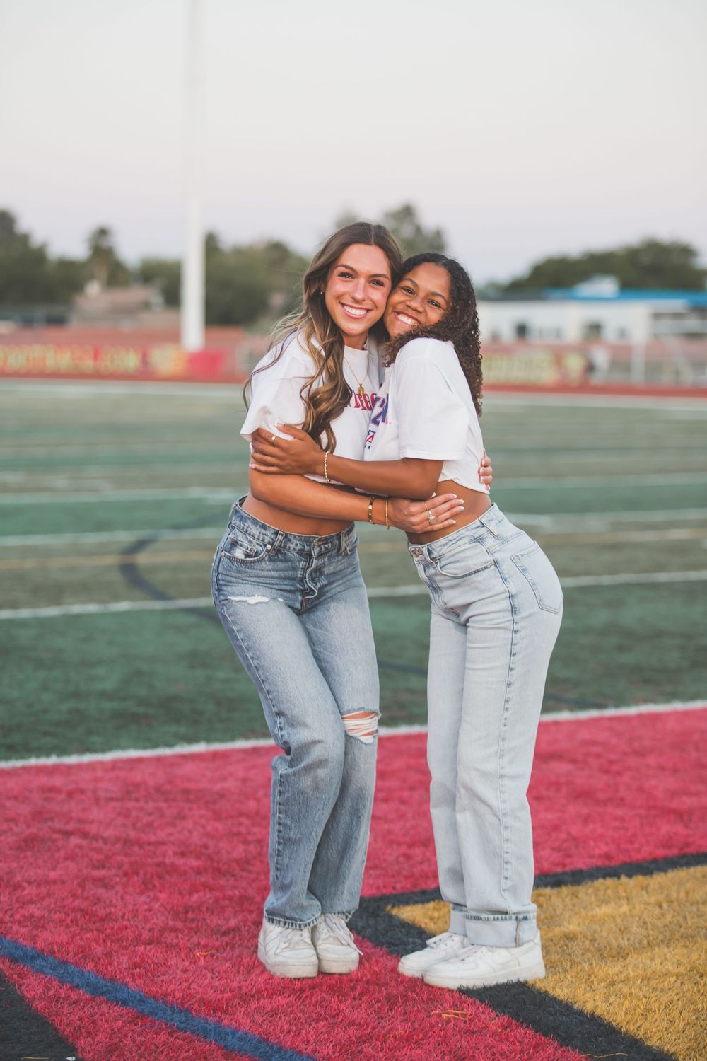 Two friends wearing white tops and jeans embrace while smiling on a sports field with colored turf markings.