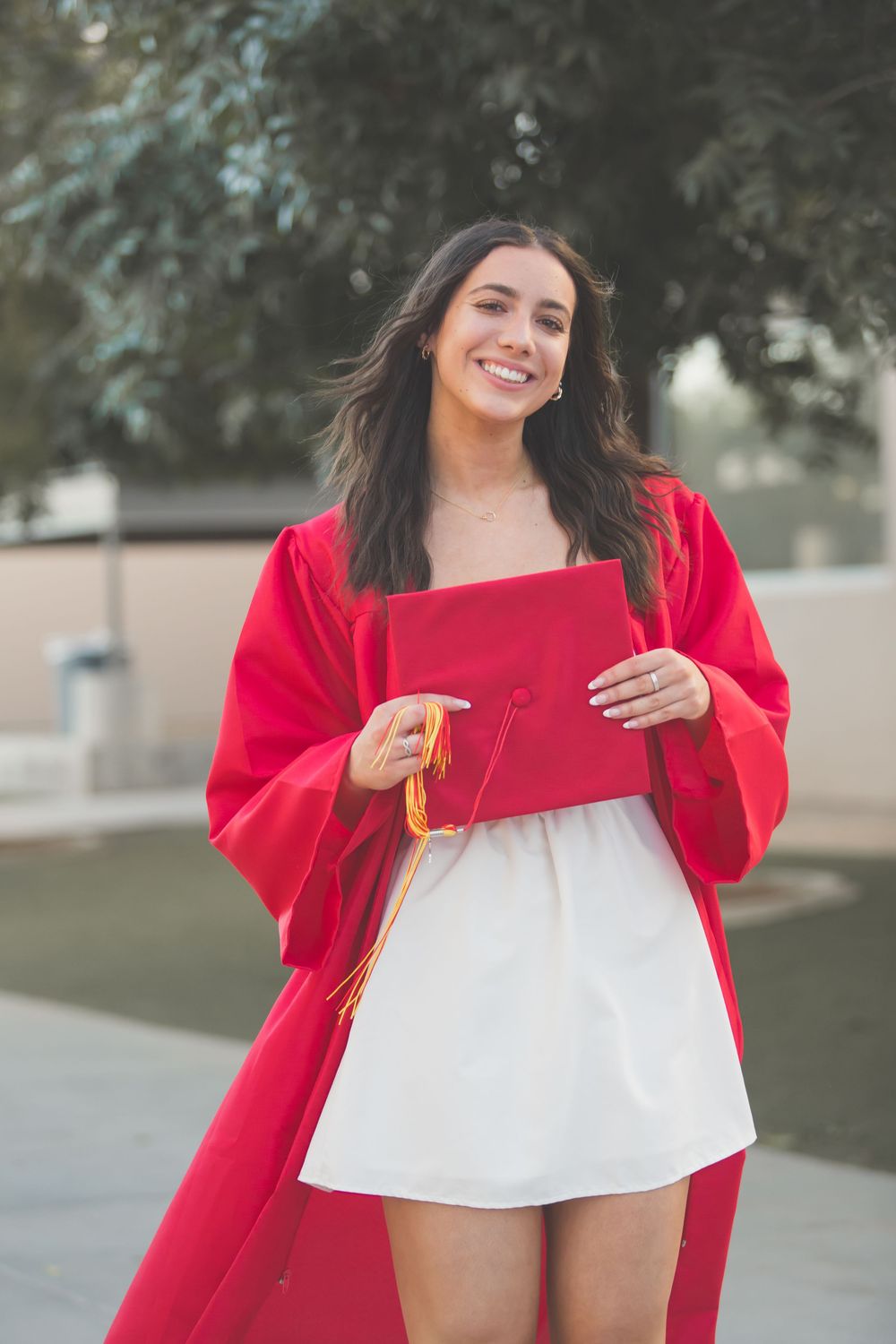 A person in a bright red coat and white dress poses playfully in an outdoor setting with trees in the background.
