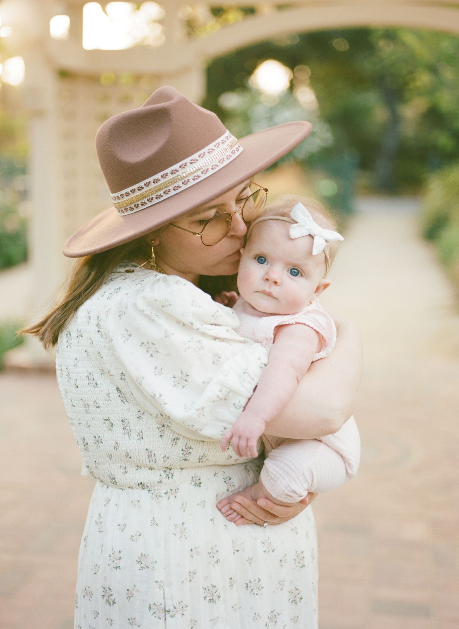 A tender moment captured outdoors showing a floral dress and brown hat against a soft bokeh background.