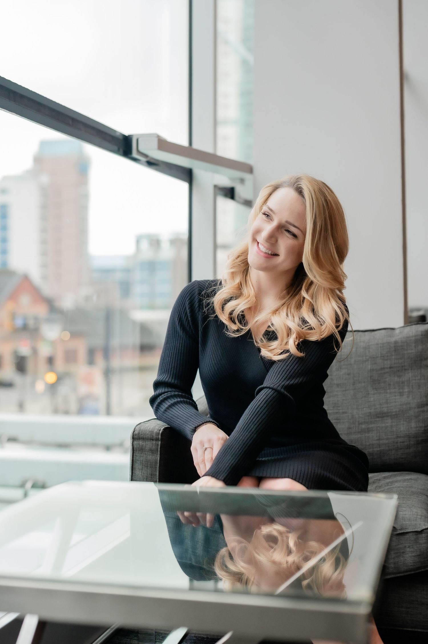 Professional woman in black sweater relaxes on gray couch near bright window in modern office setting.