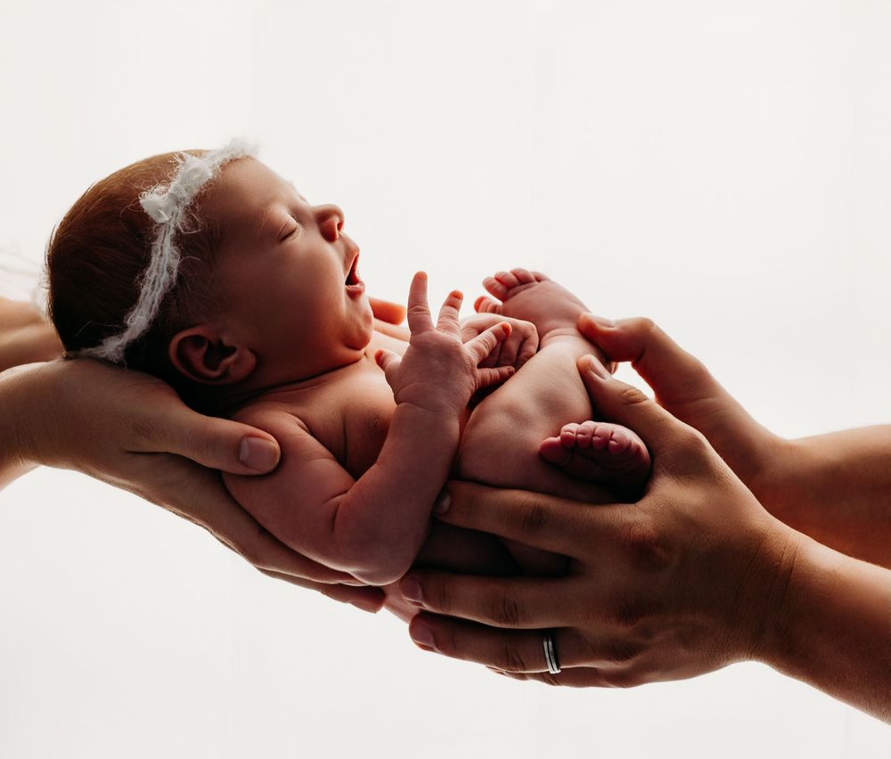 Gentle hands cradle a delicate newborn baby wearing a lace headband against a bright white background.