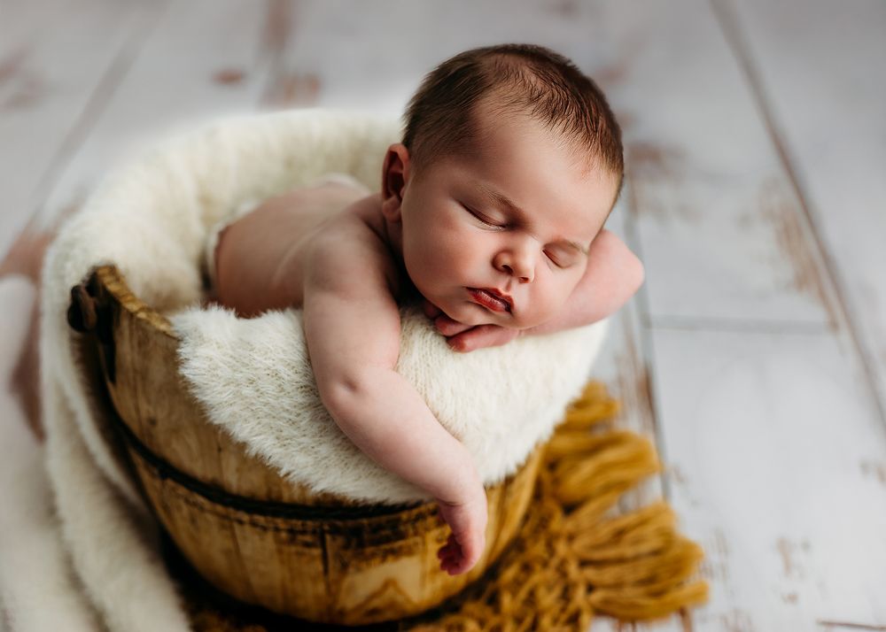 Newborn baby sleeping peacefully in round basket with white fur and mustard yellow knit blanket.