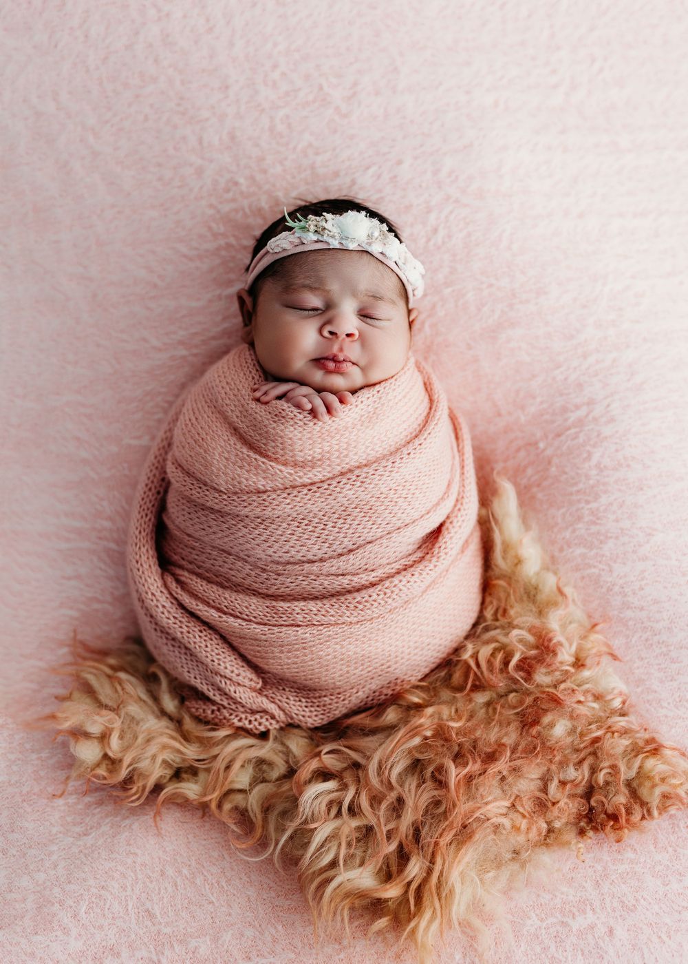 Sleeping newborn wrapped in soft pink knit blanket resting on furry beige rug with delicate lace headband.
