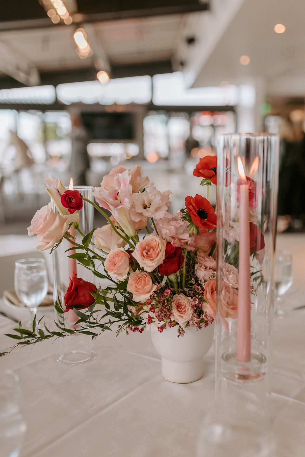 Elegant wedding centerpiece featuring pink roses and red poppies in a white vase with a tall glass cylinder vase accent.