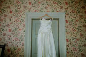 A white wedding dress hangs on a door against vintage floral wallpaper in a soft, romantic setting.