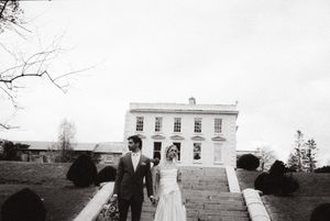 A vintage black and white photograph of a couple in formal attire walking outside a stately white mansion.