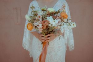 A delicate white lace wedding dress with flowing sleeves holds a peach and cream floral bouquet tied with coral ribbon.