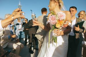 Wedding party members toast and celebrate with champagne glasses at an outdoor reception under blue sky.