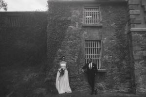 A black and white photograph of a couple standing outside an ivy-covered stone building with barred windows.