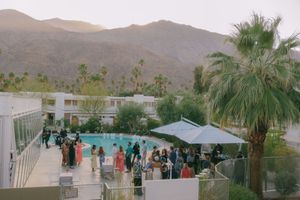 Desert resort pool area surrounded by palm trees and mountains with guests enjoying outdoor amenities at sunset.