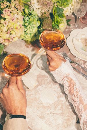 Two hands clinking rose wine glasses at an elegant floral wedding reception table with lace tablecloth.