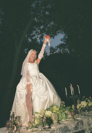 Bride in white gown raises bouquet at candlelit outdoor wedding reception table at night.