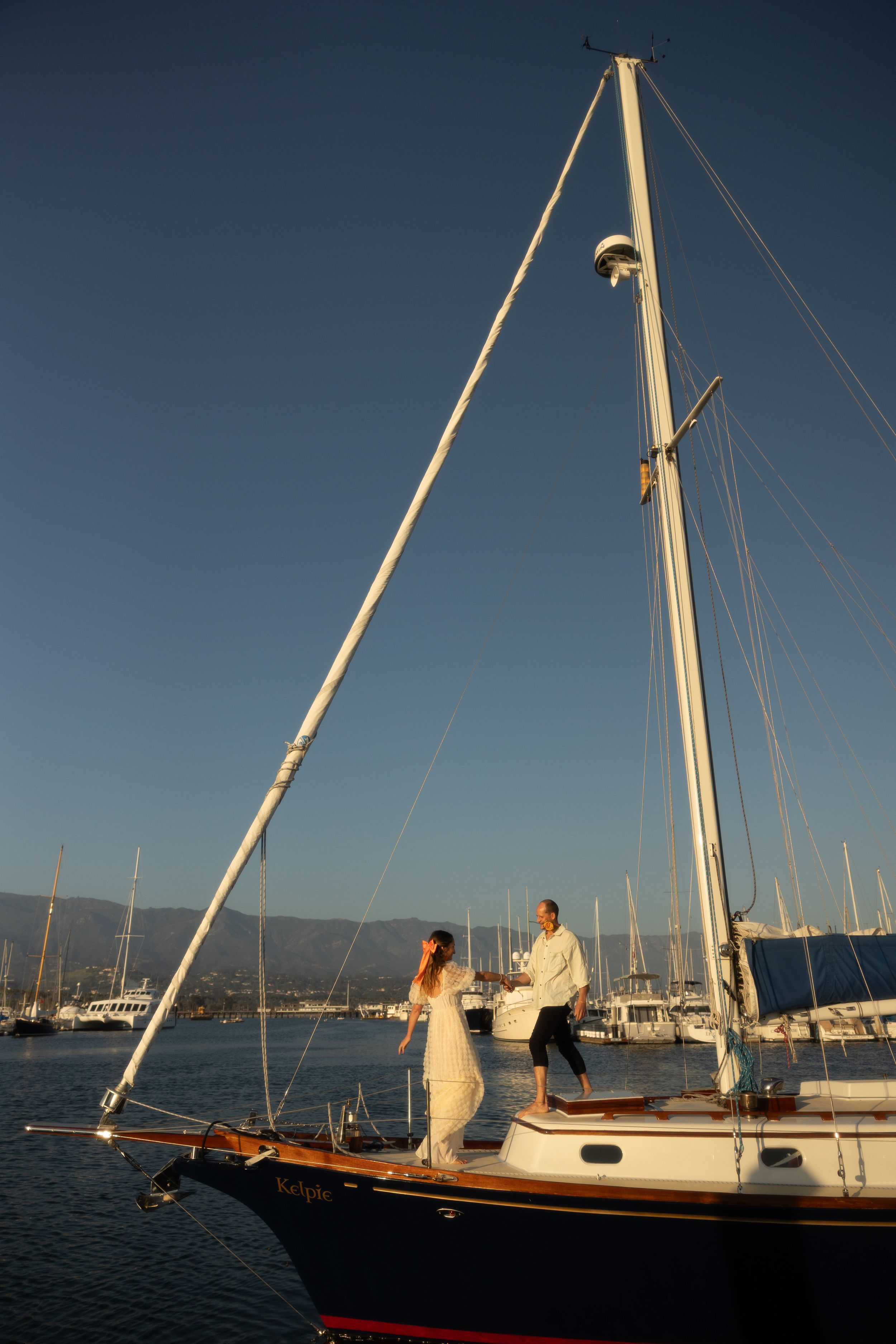 Bride and groom standing on a sailboat deck in a marina at golden hour, with masts and blue sky behind them.