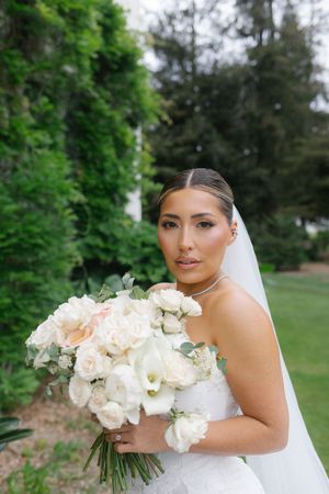 Bride in white strapless gown holding large bouquet of white and blush roses, wearing veil outdoors.