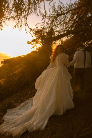 Bride in flowing white gown holds groom's hand at golden sunset on a hillside trail beneath a tree.