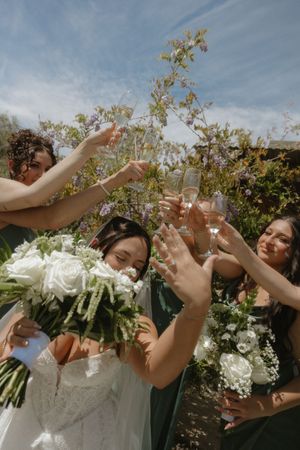 Bride and bridesmaids in green dresses toasting with champagne glasses, holding white floral bouquets outdoors.