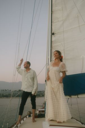 Couple standing on a sailboat at sunset, woman in white lace dress, man in light shirt, ocean and mountains behind.