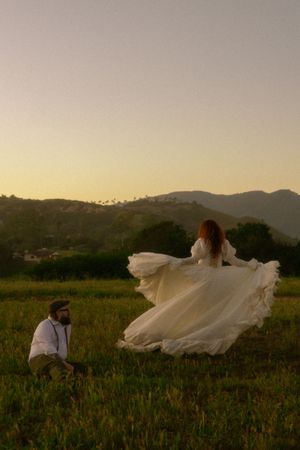 A bride in a flowing white gown twirls in a green field at sunset with mountains and a kneeling groom nearby.