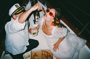 Couple in wedding attire sharing champagne over pizza on a boat, bride in white dress with red sunglasses.