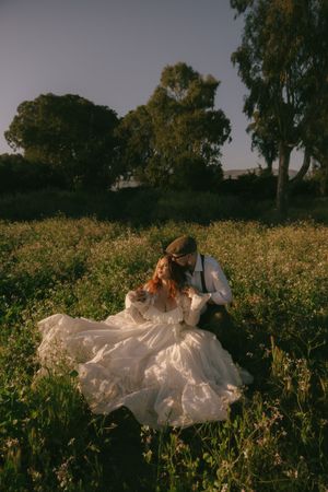 Bride in flowing white gown sits with groom in suspenders among wildflowers at golden hour.
